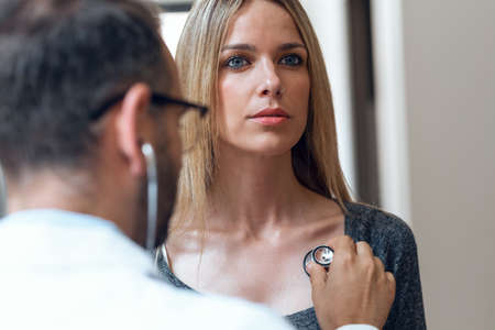 Shot of handsome mature male doctor checking beautiful young woman patient heartbeat using stethoscope in medical office.の写真素材