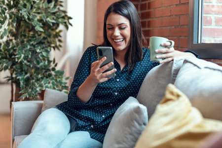 Shot of smiling woman using mobile phone while drinking a cup of coffee sitting on a couch at home.の写真素材