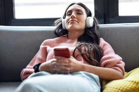 Shot of beautiful woman listening to music while using smartphone sitting on sofa at home.の写真素材
