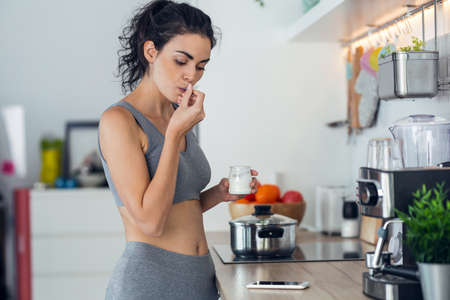 Shot of sporty young sporty woman eating iogurt while using mobile phone standing in the kitchen at home.の写真素材