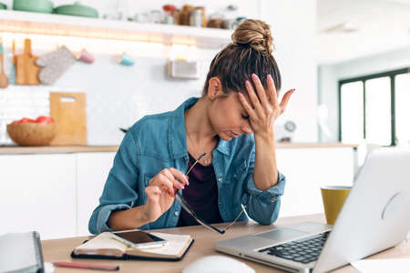 Shot of stressed business woman working from home on laptop looking worried, tired and overwhelmed int he kitchen at home.の写真素材