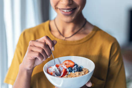 Close-up of beautiful mature woman eating cereals and fruits while standing next to the window at home.の写真素材