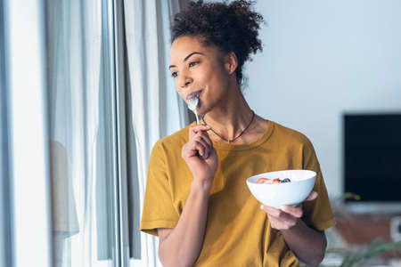 Shot of beautiful mature woman eating cereals and fruits while standing next to the window at home.の写真素材