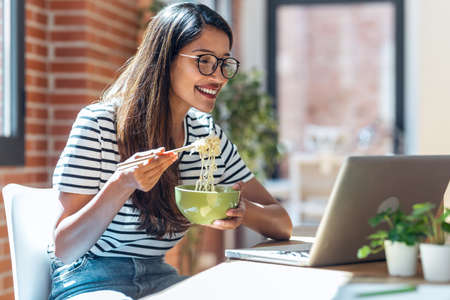 Shot of happy woman eating noodles with chopsticks while doing a videocall with laptop in living...