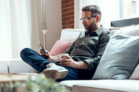 Shot of mature male using smartphone texting while drinking a cup of coffee and relaxing on sofa at home.の写真素材