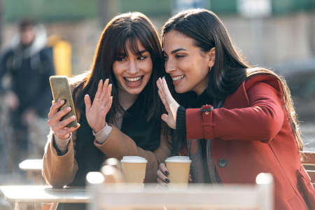 Shot of two attractive friends enjoying coffee together while doing a videocall with smartphone sitting on the terrace of a bar.の写真素材