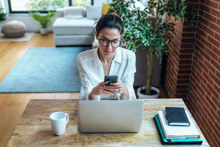 Shot of smiling business woman using her mobile phone while working with computer in the office.の写真素材