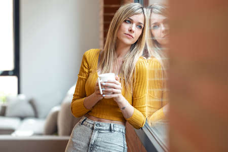 Shot of beautiful young woman drinking a cup of coffee while looking forward standing next to the window at home.の写真素材