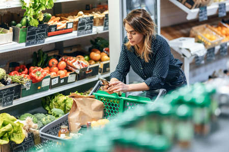 Shot of beauty woman walking with shopping cart while taking products from shelf at the groceryの写真素材