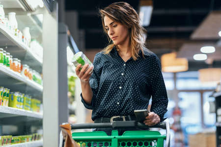 Shot of beauty woman walking with shopping cart while taking products from shelf at the grocery