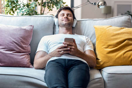 Shot of handsome man listening to music with headphone while using smartphone lying on sofa at home.の写真素材