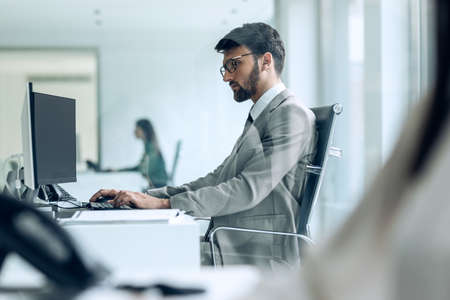 Shot of handsome young entrepreneur working with computer while reading some documents in modern startup office.の写真素材