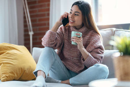Shot of pretty woman using mobile phone while drinking cup of coffee on sofa at home.の写真素材