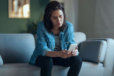 Shot of depressed woman using smartphone while sitting on the coach at home