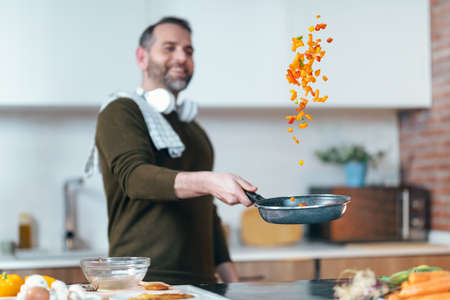 Shot of handsome mature man sautÃ©ing vegetables in the pan while having fun in the kitchen at home.の写真素材