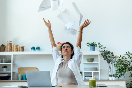 Shot of worried woman tossing invoices and documents into the air while working on the laptop at home.の写真素材
