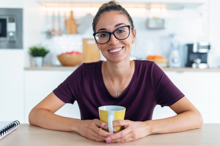 Shot of smiling young woman waving through the laptop web camera while holding a cup of coffee in the kitchen at home.の写真素材
