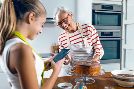 Shot of cute grandmother taking photos with her smartphone while grandmother making tasty chocolate cake in the kitchen.の写真素材