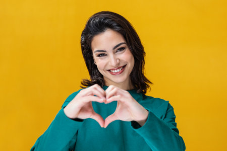 Portrait of romantic happy woman making heart shape with hands while smiling to camera isolated on yellowの写真素材