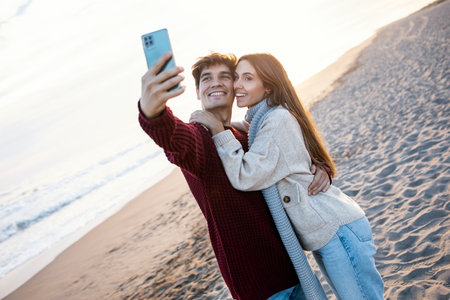 Shot of beautiful young couple making photos with mobile phone in a cold winter on the beach.の写真素材
