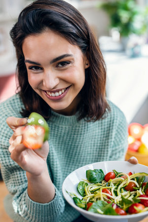Shot of beautiful smiling woman eating healthy salad while sitting on the table at home.の写真素材