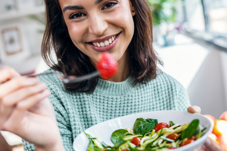 Shot of beautiful smiling woman eating healthy salad while sitting on the table at home.の写真素材