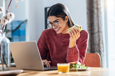 Shot of beautiful young woman working with laptop while having healthy sandwich for breakfast at homeの写真素材