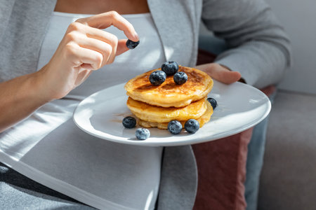 Shot of beautiful happy pregnant woman in panties eating pancakes with red fruits lying on sofa at homeの写真素材