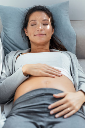 Shot of beautiful pregnant woman touching her belly while relaxing lying on sofa at homeの写真素材