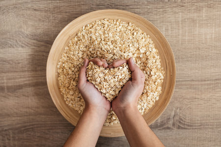 Close up of woman holding heart-shaped hands of oatmeal from a wooden bowl in the kitchen at homeの写真素材