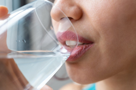 Close up of beautiful sporty woman drinking a glass of water while standing near the window at homeの写真素材