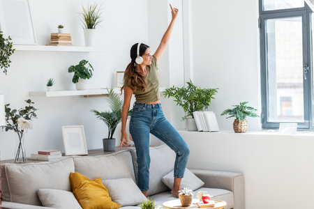 Shot of motivated happy woman singing and dancing on couch while listening to music with headphones at home.の写真素材