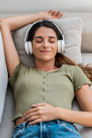 Shot of beautiful kind woman relaxing while listening to music with headphones lying on couch at...
