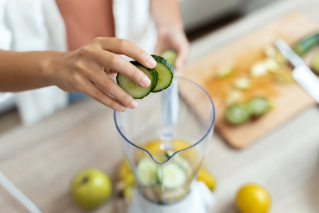 Close up of pretty young woman putting different kinds of vegetables and fruits in to the blender for preparing detox juice in the kitchen at homeの写真素材
