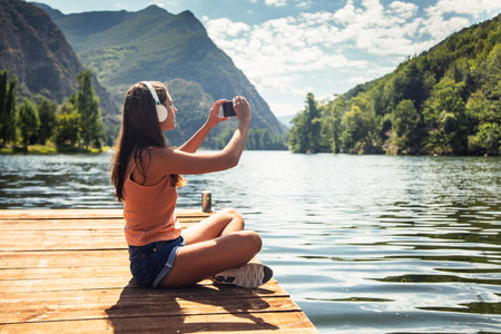 Shot of happy woman listening to music with headphones while taking photos with smartphone to the lakeの写真素材
