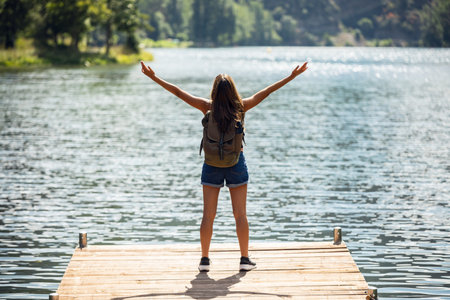 Shot of back view of young woman relaxing and raising her arms while staying near the lake in a mountain landscapeの写真素材