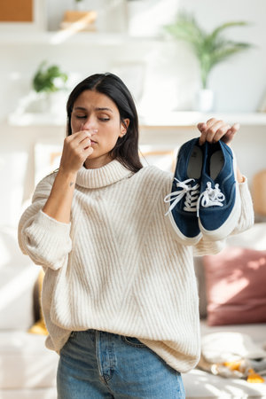 Shot of disgusted beautiful woman holding stinky shoes in the living room at homeの写真素材