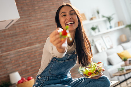 Portrait of beautiful smiling woman eating healthy salad while sitting on the kitchen table at home.の写真素材