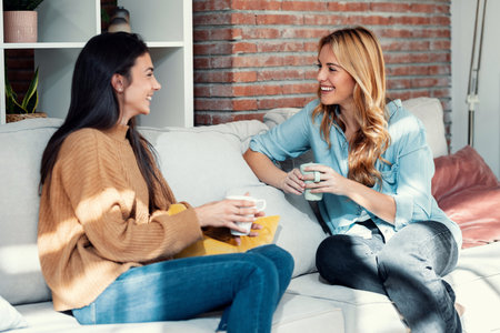 Shot of two smiling young women talking while drinking coffee sitting on couch in the living room at home.の写真素材