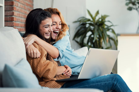 Shot of two happy beautiful women doing a video call with laptop while hugging each other sitting on the couch at home.の写真素材
