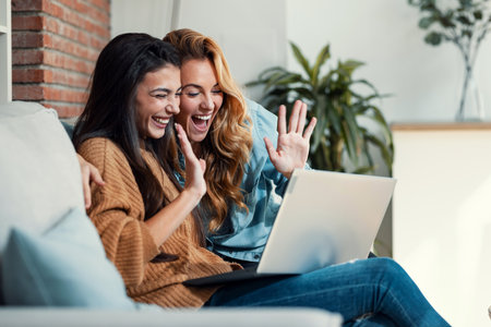 Shot of two happy beautiful women doing a video call with laptop while waving hand sitting on the couch at home.の写真素材