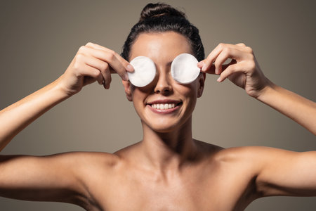 Portrait of funny young woman covering eyes with make-up pads while smiling on isolated brown backgroundの写真素材