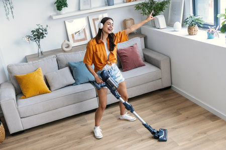 Shot of young happy woman listening and dancing to music while cleaning the living room floor with a vacuum cleanerの写真素材