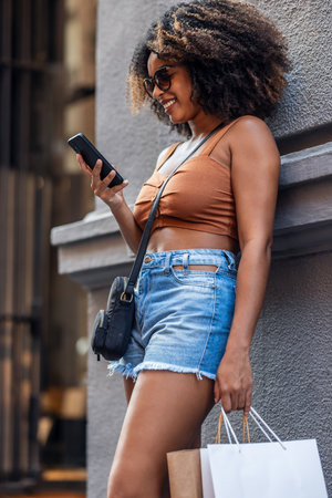 Shot of happy woman using smartphone while carrying shopping bags leaning against the wall in the cityの写真素材