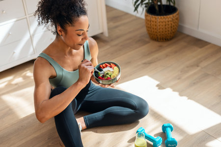 Shot of athletic woman eating a healthy fruit bowl while sitting on floor in the kitchen at homeの写真素材