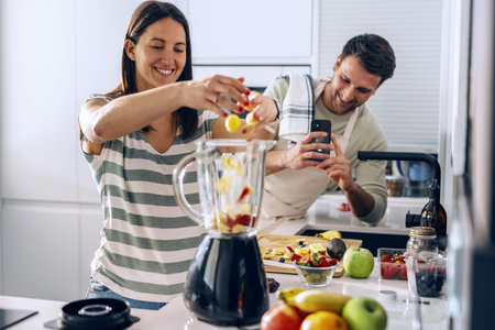 Shot of pretty woman preparing detox juice with blender while her husband recording with smartphone...