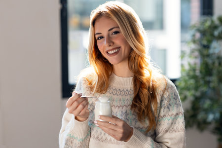 Shot of happy beautiful woman eating yogurt while standing in living room at home