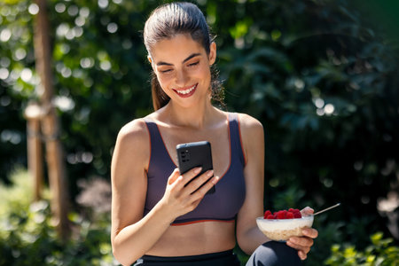 Shot of attractive woman eating healthy breakfast while using her smartphone in the gardenの写真素材
