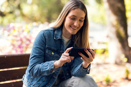 Shot of pretty young woman using her mobile phone while sitting on a bench in the park.の写真素材