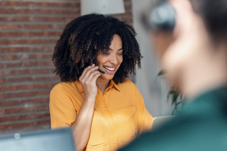 Shot of business woman making video call with computer while talking with earphone sitting in modern startup officeの写真素材
